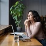 Young dreamy smiling female entrepreneur thinking about business goals and objectives, enjoying good window view while working in office, pleased businesswoman resting from computer work